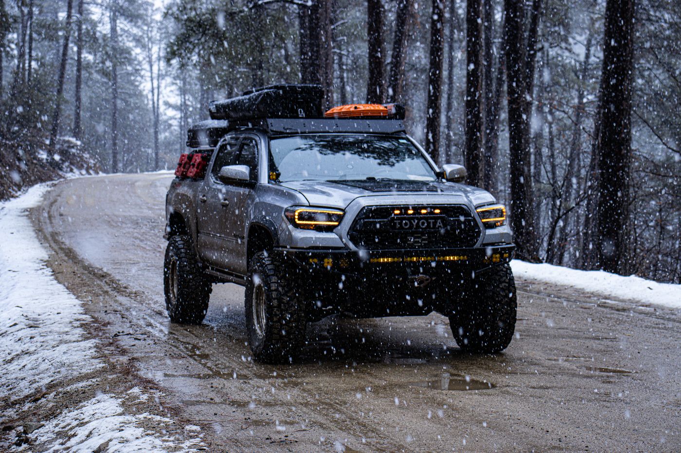 Tacoma driving through falling snow on a forest road, headlights on